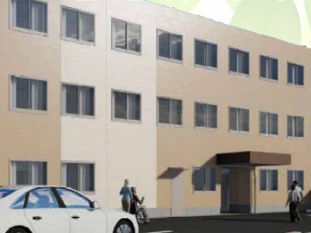 ブラウン系の外観で3階建ての建物がある敷地内には駐車場があり、車が止まっている。車いすの人と付き添いの人もいる。