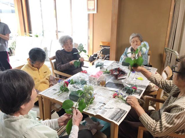 新聞紙を広げたテーブルを囲んでフラワーアレンジメントを楽しんでいる高齢女性たち。共有スペースには大きな窓が付いている。