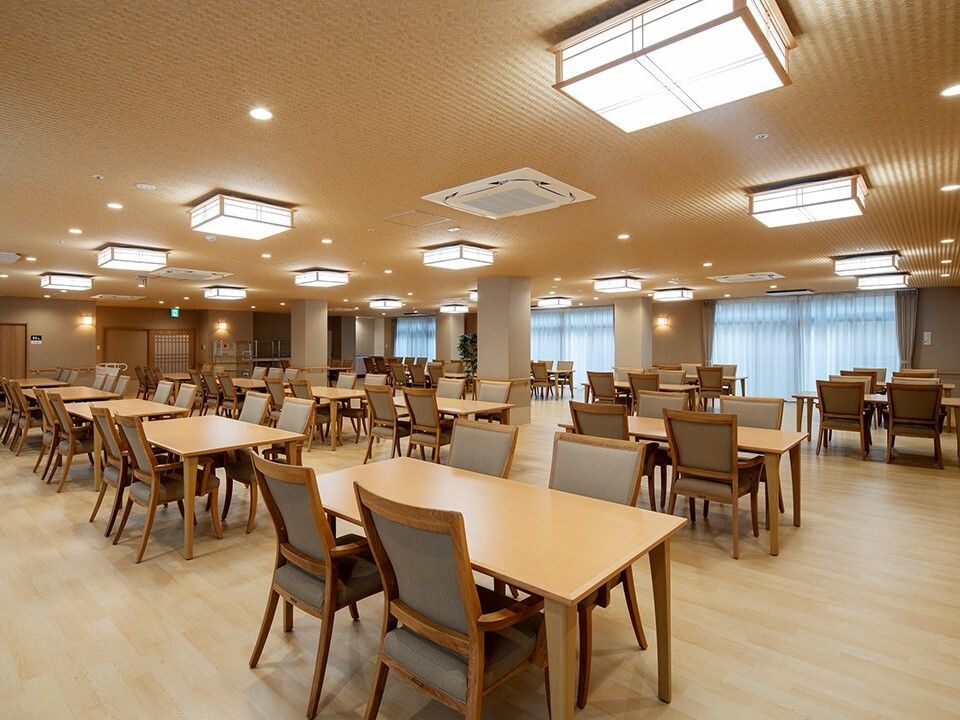 木製のダイニングテーブルが複数置かれている食堂にはレースカーテンとカーテンが付いたおおきな窓がある。