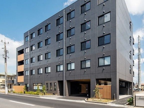 グレー系の外観の5階建ての建物は日当たりのいい場所に建っている。周りには草木が植えられている。