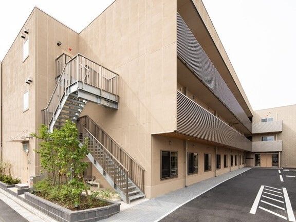 各フロアにベランダが付いたベージュの外壁の3階建ての建物の側面には屋外階段も付いている。