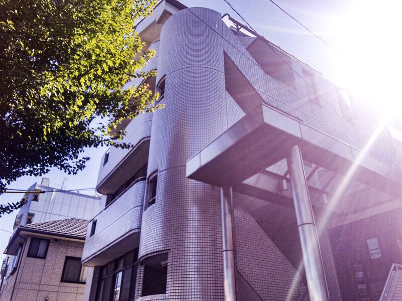 日当たりのいい場所に建っているグレー系の建物の前には大きな木が植えられている。各フロアにベランダもある。