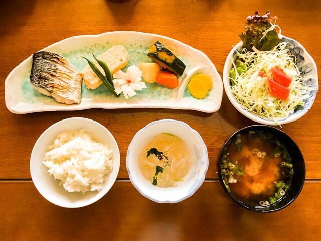 サバの塩焼きや卵焼き、煮物、漬物、サラダ、みそ汁、野菜の和え物などが並び、白いご飯もある。