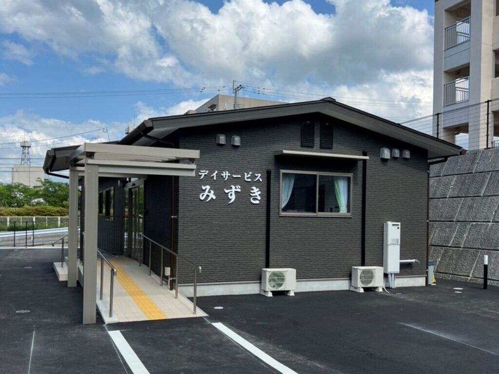 デイサービスの名前が白い文字で書かれたダークグレーの外壁の平屋建ての建物。玄関先には手すり付きのスロープがある。