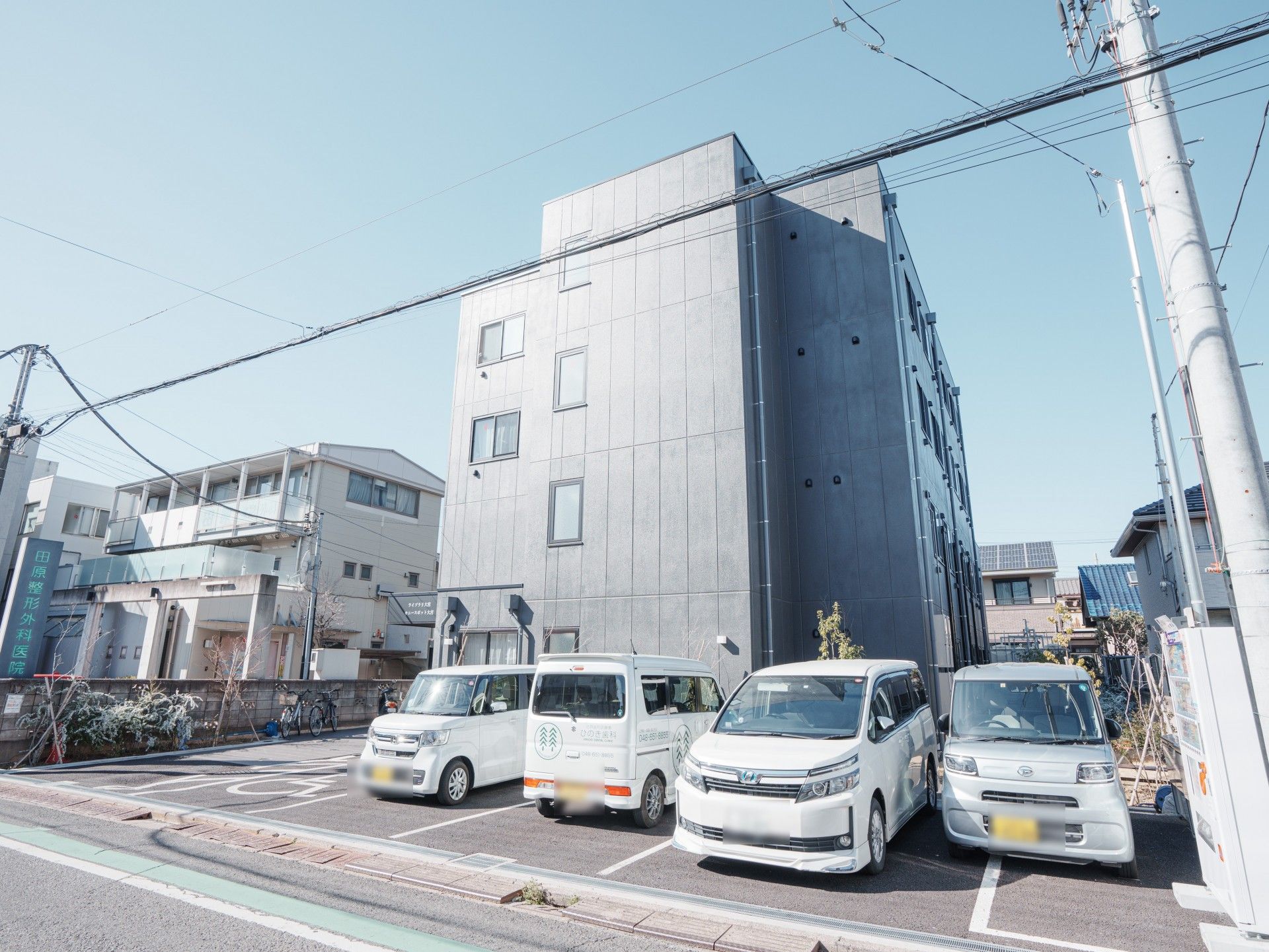 車いす対応の駐車場スペースなどが建物の前にあり、草木が植えられている。ダークグレーの４階建ての建物。