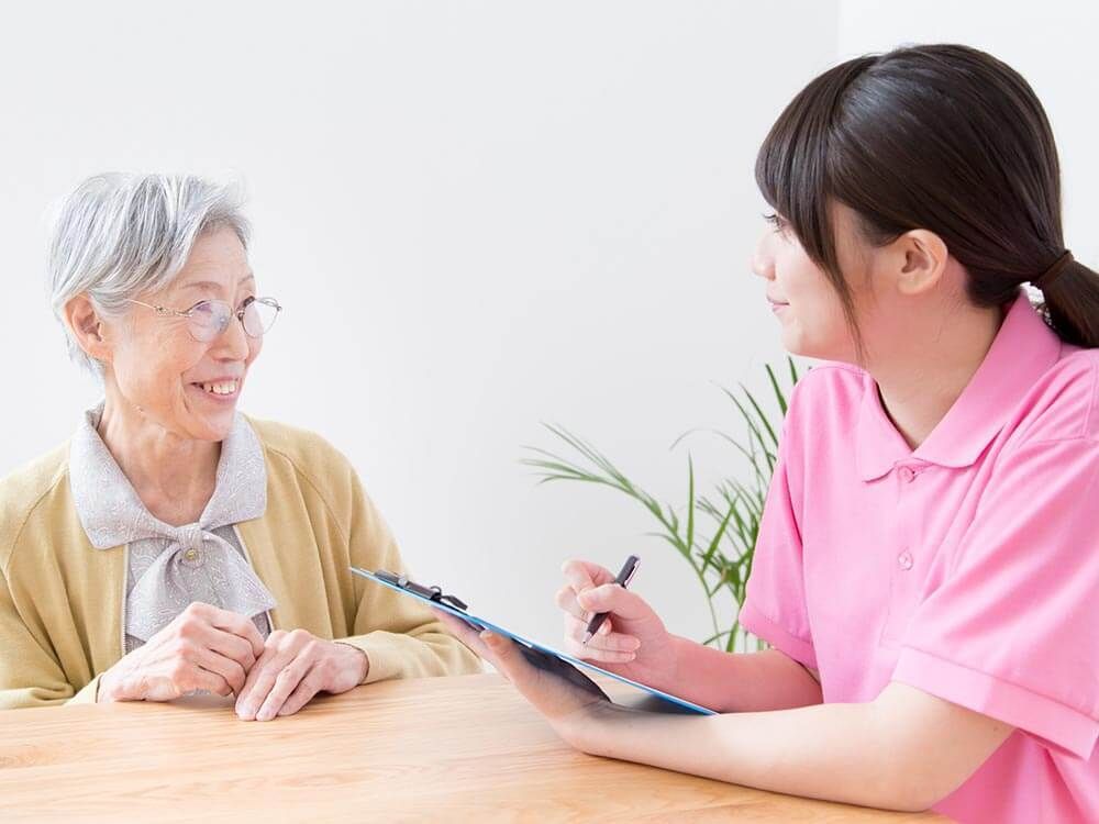 眼鏡をかけた高齢女性とポロシャツを着た女性スタッフがテーブルを囲んで笑顔で話している。