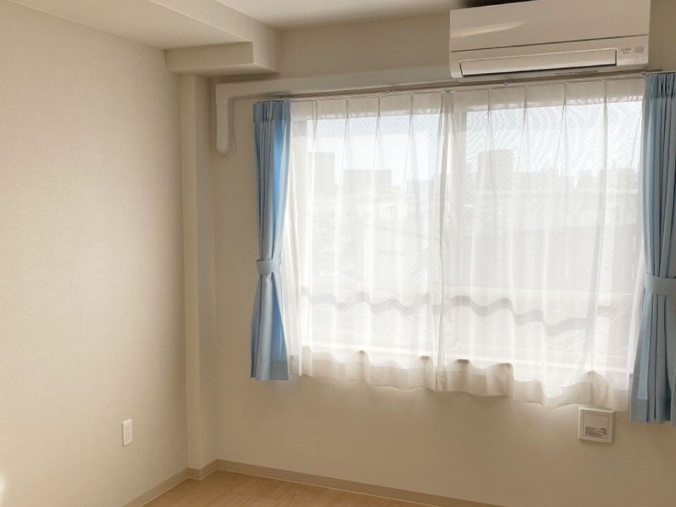 エアコンが付いたフローリングの部屋。レースカーテンとカーテンが付いたおおきな窓があり、家具は置かれていない。