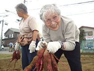 大きなサツマイモを持って笑顔になっている軍手をした眼鏡をかけた高齢女性。畑は広く、周りに民家がある。