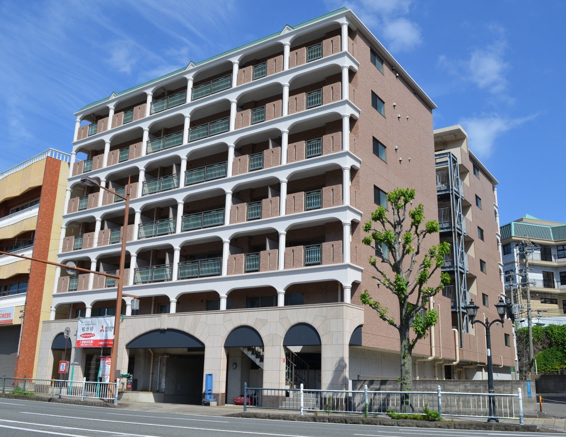 ブラウン系の7階建ての建物は各フロアにベランダがあり、周りには草木も植えられ、上空には電線が多くある。