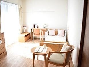 寝具付きのベッドやタンス、デスクセットやテーブルセットが置かれているフローリングの部屋には大きな窓があり明るい。