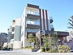 グレー系の大きな建物の玄関には車寄せが付き、建物の周りには草木が植えられている。
