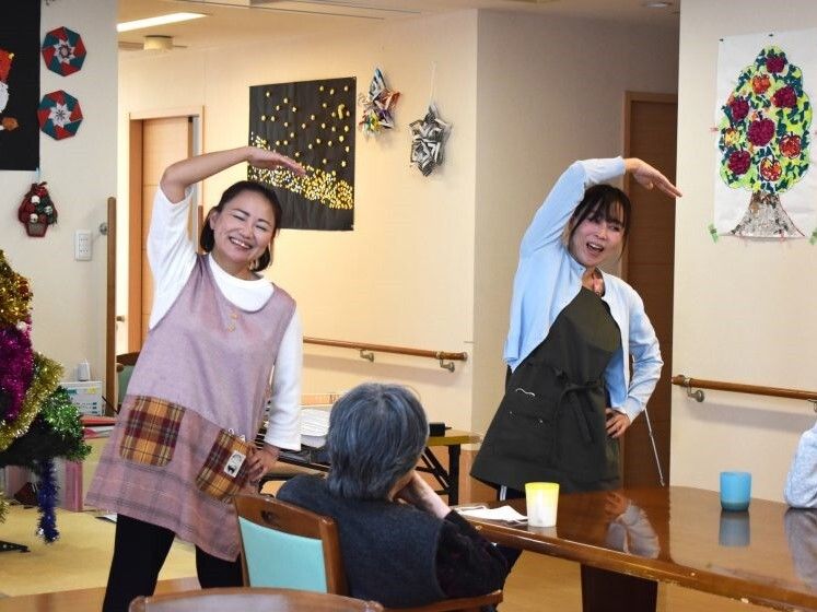 元気よく体操をしている女性スタッフ２名。高齢女性たちがダイニングテーブルを囲んでいる。