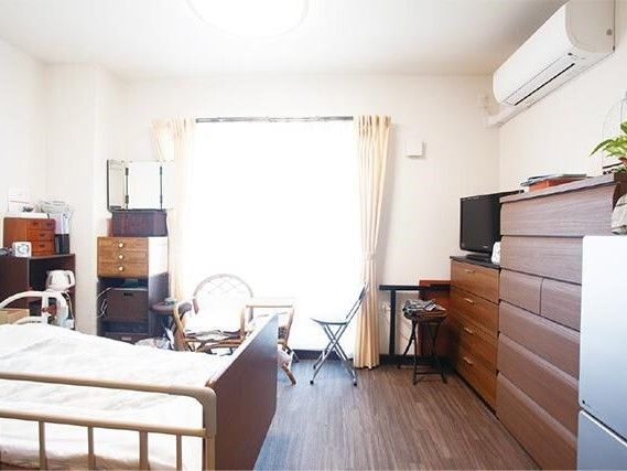 フローリングの部屋には介護ベッドやタンス、椅子、テーブル、エアコン、冷蔵庫、テレビなどが置かれている。