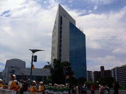 斜め屋根の佇まいが印象的な高層ビルが建っている。手前の広場では、子どもたちが楽器を演奏する姿が見られる。