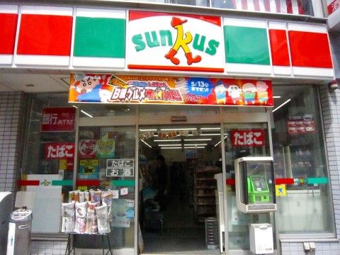 赤色と緑色のコントラストが印象的なコンビニがある。出入口のところに朝刊が置かれている。公衆電話もある