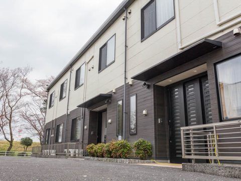 白とダークグレーの外壁の２階建ての建物は玄関に屋根があり、周りに草木が植えられている。