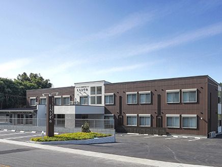 青空の下に建っている２階建ての建物の前には駐車場があり、周りには緑が多い。
