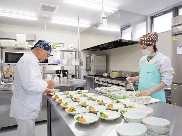 ステンレス製の本格的な調理器具がそろった厨房で男女２名のスタッフが作業をしている。