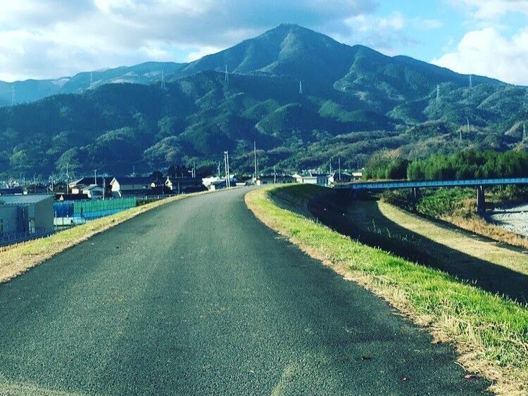 緑あふれる自然豊かな環境。右に川、目の前に大きな山、左側に民家、真ん中に道路がある。