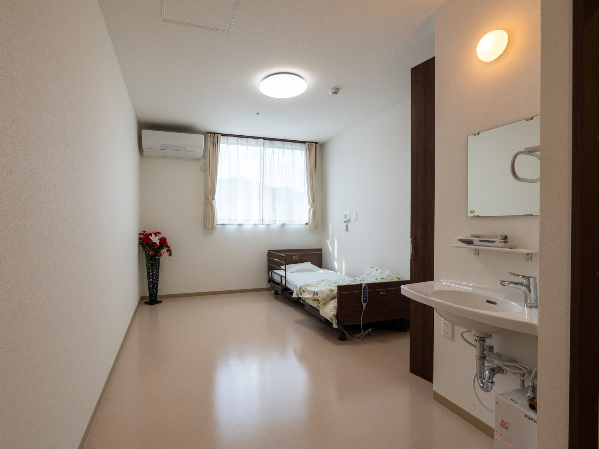 レースカーテンとカーテンが付いた窓がある部屋には寝具付きのベッドや観葉植物があり、洗面所も付いている。