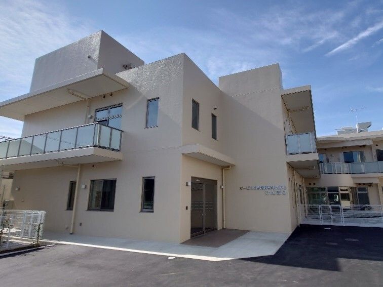 白い外壁の２階建ての建物は玄関にマットが敷かれ、２階部分にはベランダがあり、外壁には住宅名が書かれている。