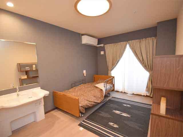 大きな窓にはカーテンが付き、フローリングの部屋には車いす対応の洗面所や寝具付きベッドがある。