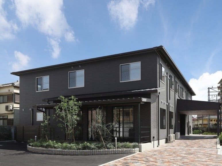三角屋根が付いたダークグレーの外壁の２階建ての建物の周りには草木が植えられ、日当たり良好。
