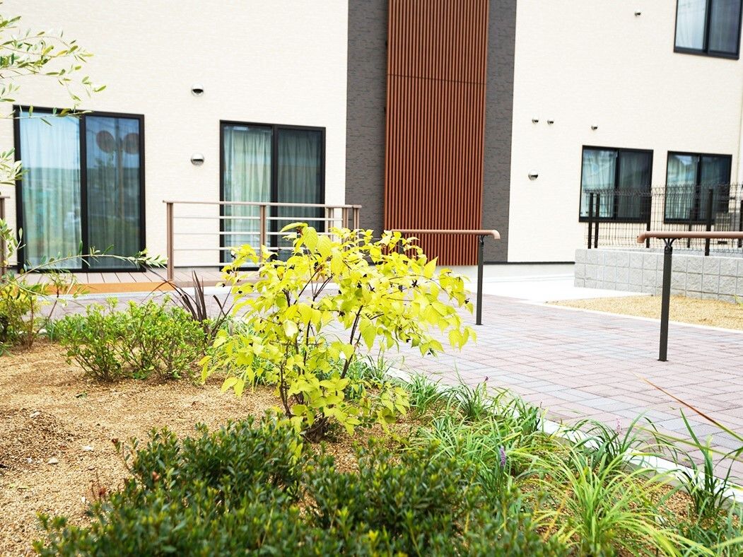 敷地内には手すりが設置され、ベージュの外観の２階建ての建物の周りには草木が植えられた庭もある。