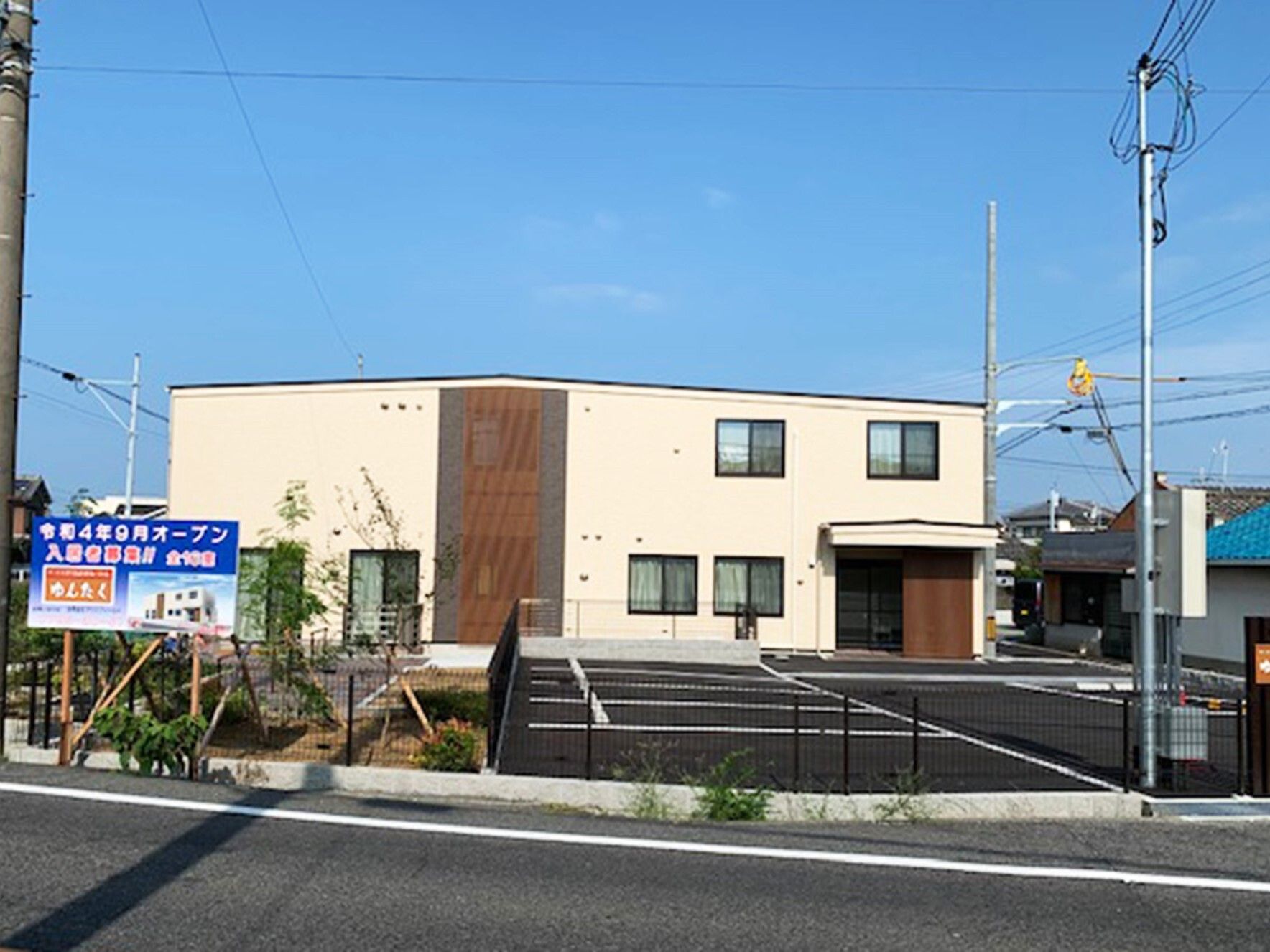 青い空の下に建っているベージュの外観の２階建ての建物の周りには草木が植えられた庭があり、駐車場も完備されている。