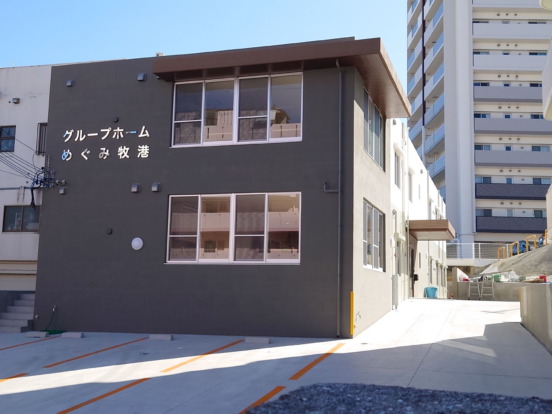 ２階建ての建物は横に長く、白とグレーの外壁で、玄関周りに段差はなく、屋根が付いている。