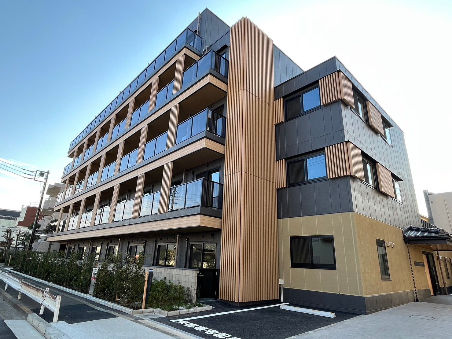 ベージュとダークグレーの外壁の５階建ての建物の各フロアにベランダがあり、駐車スペースも付いている。