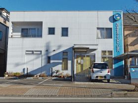 白を基調とした２階建ての建物は日当たり良好で玄関先には屋根があり、駐車場もある。