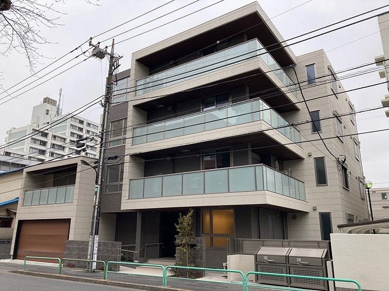 4階建ての建物は各フロアにベランダがあり、周りにはたくさんの建物が立っていて、玄関先には木が植えられている。