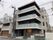 4階建ての建物は各フロアにベランダがあり、周りにはたくさんの建物が立っていて、玄関先には木が植えられている。