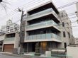 4階建ての建物は各フロアにベランダがあり、周りにはたくさんの建物が立っていて、玄関先には木が植えられている。