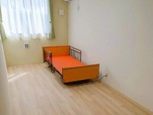 レースカーテンとカーテンが付いた大きな窓が付いているフローリングの部屋には介護ベッドが置かれている。