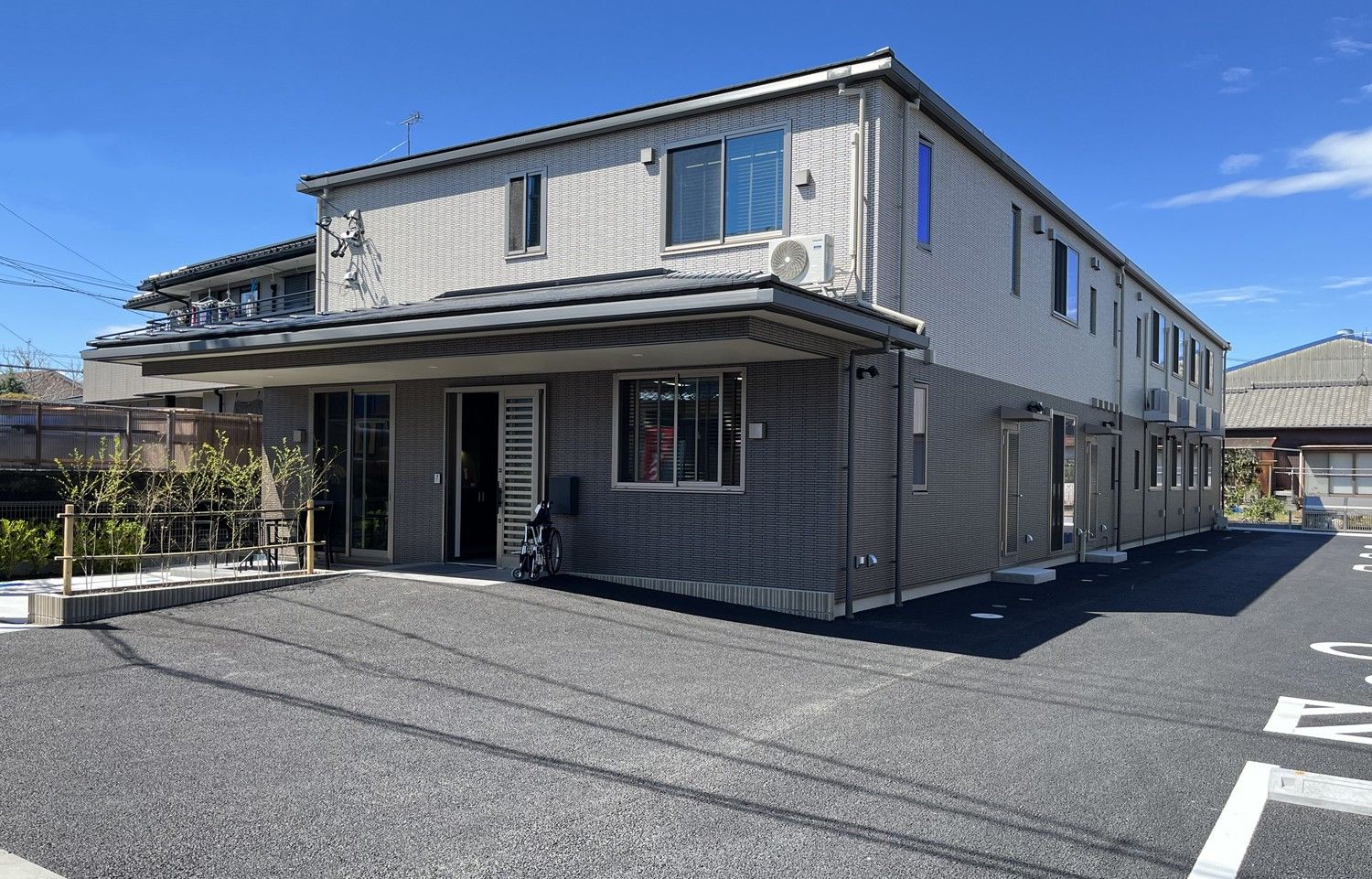 ベージュの外壁の２階建ての建物の敷地内には広い駐車場や小さな菜園があり、日当たりのいい場所に建っている。