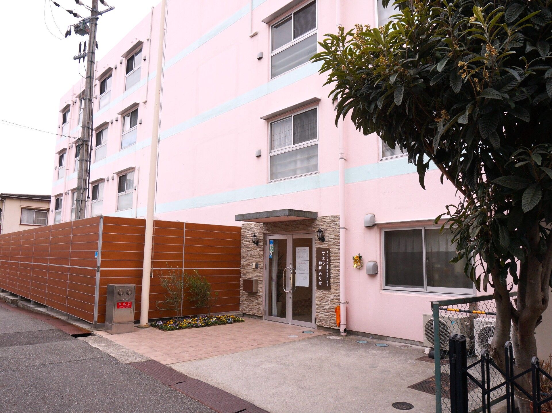 屋外で笑顔で散歩をしているデニムシャツを着た車いすの高齢女性とエプロンを付けた女性スタッフ。