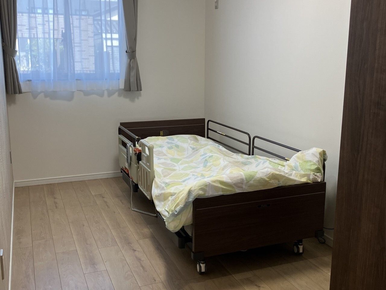 レースカーテンとカーテンが付いた窓があるフローリングの部屋には寝具付きベッドが置かれている。