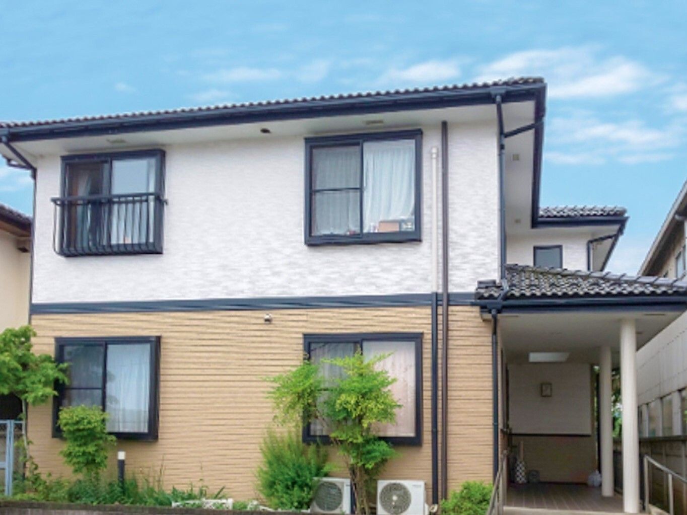 ２階建ての建物は日当たり良好で玄関先には屋根があり、建物の周りには草木が植えられている。