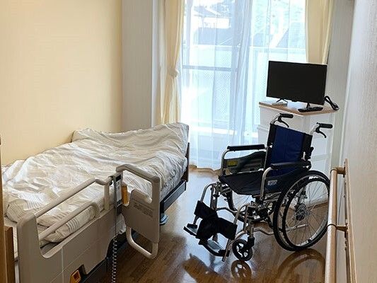 レースカーテンとカーテンが付いた大きな窓があるフローリングの部屋には寝具付きのベッドが置かれ、車いすやテレビもある。