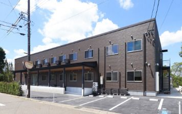 ブラウン系の2階建ての建物の前伊には駐車場があり、建物の側面には屋外階段も設置されている。
