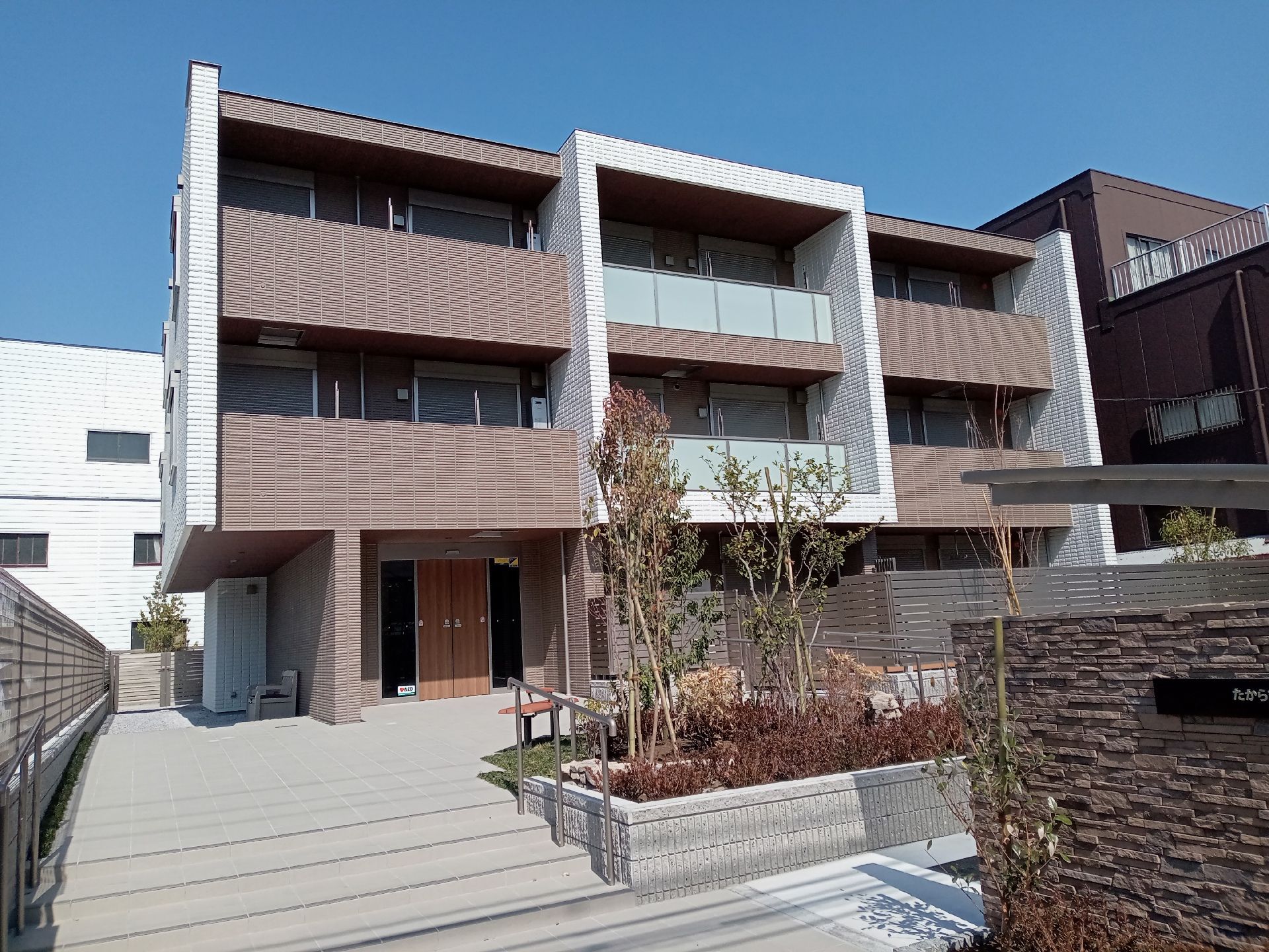 3階建ての建物の各フロアにベランダがあり、敷地内には草木が植えられたスペースもあり、階段には手すりがある。
