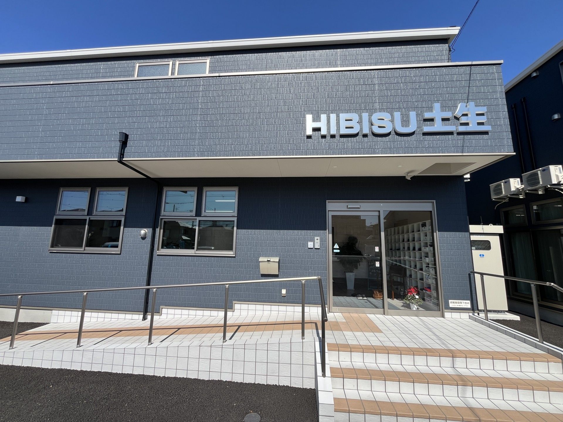 赤い車が2階建ての建物の前に泊まり、自転車で行き飼う人や車いすを使用している人がいる。