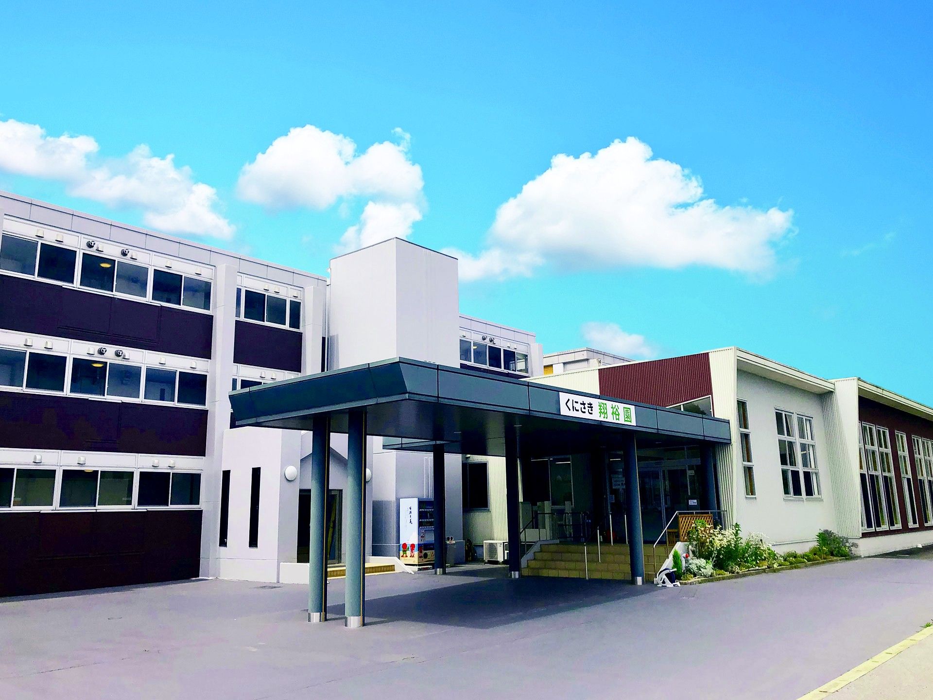 青空の下に建っている3階建ての建物の玄関には車寄せが付いている。