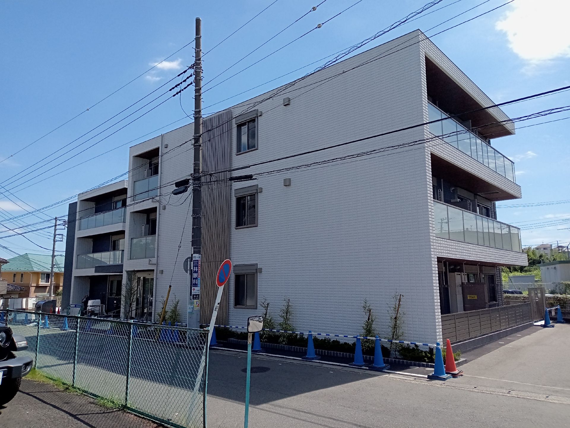 白を基調とした3階建ての建物の各フロアにベランダがあり、草木が植えられている。道路に面していて周りに住宅もある。