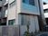 グレーの外壁建物は日当たりのいい場所に建っている。門があり、その周りには草木が植えられている。