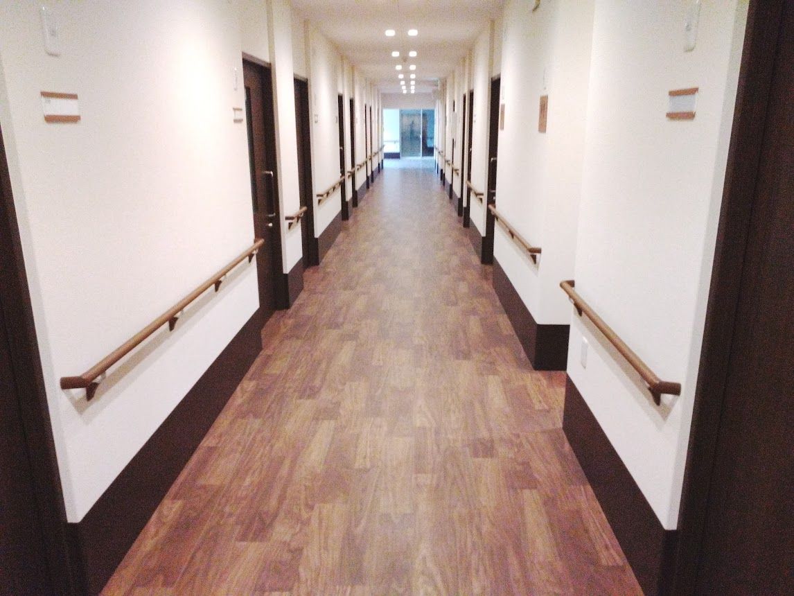 緑の多い屋外で誰かに手を振っている車いすの高齢女性と付き添いの男性スタッフ。