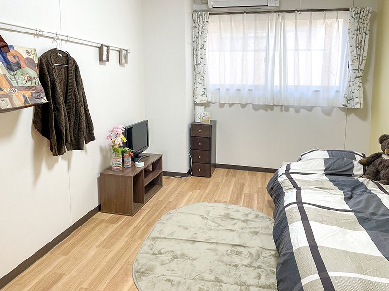 レースカーテンとカーテンが付いた大きな窓があるフローリングの部屋には寝具付きのベッドや棚がある。