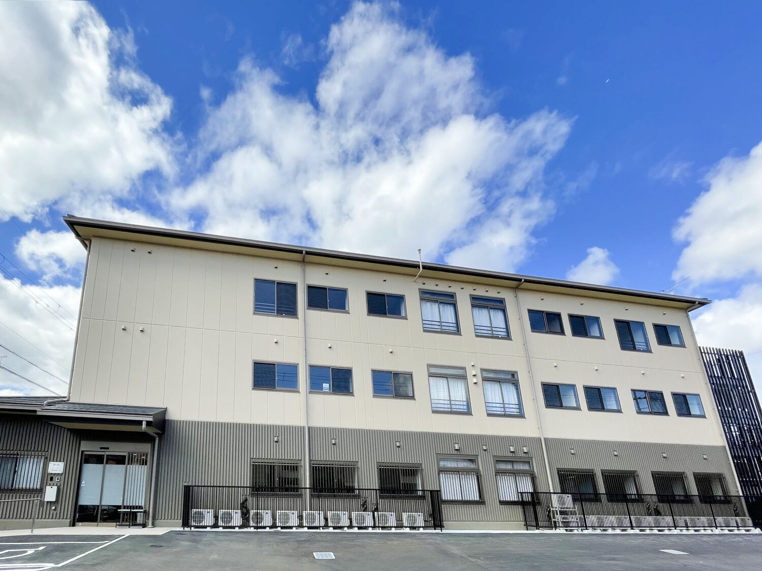 青い空の下に建っている3階建ての建物の前には駐車場があり、玄関には屋根が付いている。